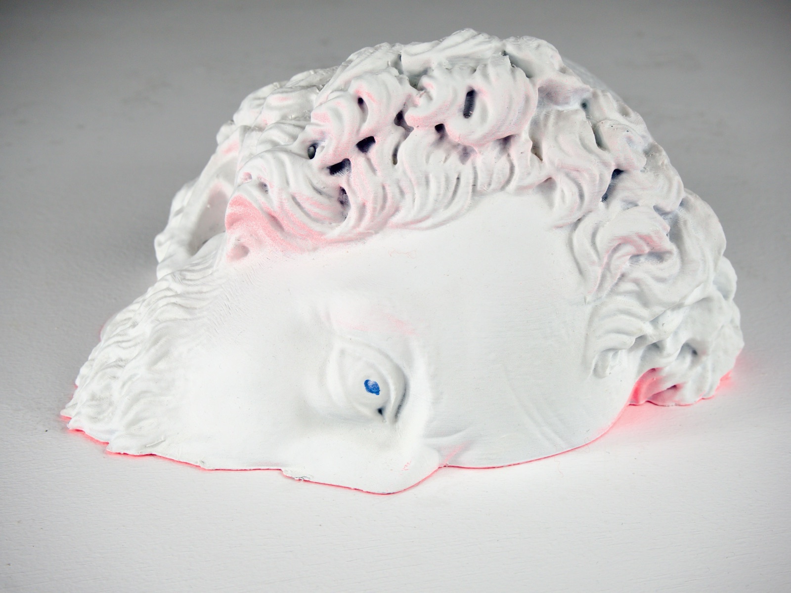 Tête imprimée en 3D posée sur le côté : surface blanche au gesso, cheveux bouclés, yeux bleus, lueur rose fluorescente sur les bords