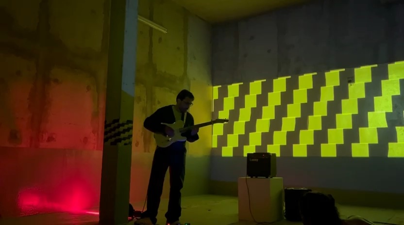 Guillaume Milochau à la guitare électrique dans un sous-sol en béton, devant une projection de damier jaune